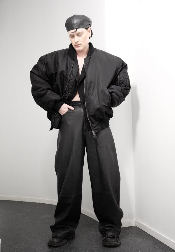 Persona, kas valkā pārizmēra melnu bumbvedēja jakas, platu ādas bikses, melnu baretu un melnas kurpes, stāv pret baltu stūra sienu.