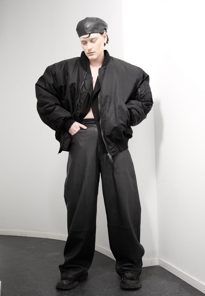 Personne portant une veste bomber noire surdimensionnée, un pantalon large en cuir, un béret noir et des chaussures noires, se tenant contre un mur d'angle blanc.