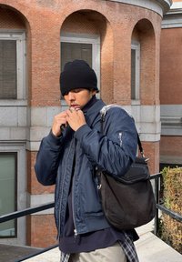 Ναυτική bomber jacket με ribbed μανσέτες, μαύρο beanie και μια μαύρη χαλαρή τσάντα ώμου. Διακριτή υφή και φερμουάρ στην καμπαρντίνα.