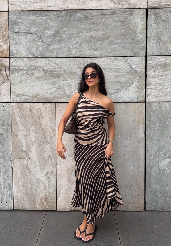Donna con occhiali da sole e abito monospalla con stampa a zebra, che tiene una borsa, in piedi davanti a una grande parete di mattonelle di pietra.