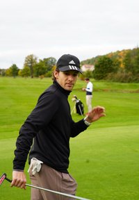 Športnik v črni majici z dolgimi rokavi in črni kapici s tremi belimi črtami, drži golf palico, ozadje iz zelene trave.