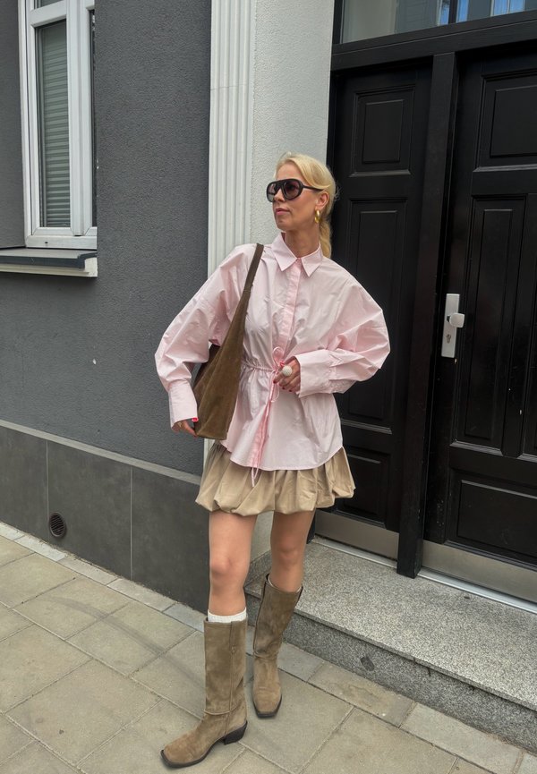 Femme portant une chemise rose oversize, une jupe beige, des bottes marron montantes et des lunettes de soleil, debout sur le trottoir près d'une porte noire avec un sac à bandoulière marron.