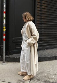 Personne aux cheveux bruns courts portant un long manteau beige, un pull gris, un pantalon blanc et des chaussures beige sans lacets debout sur un trottoir en ville.