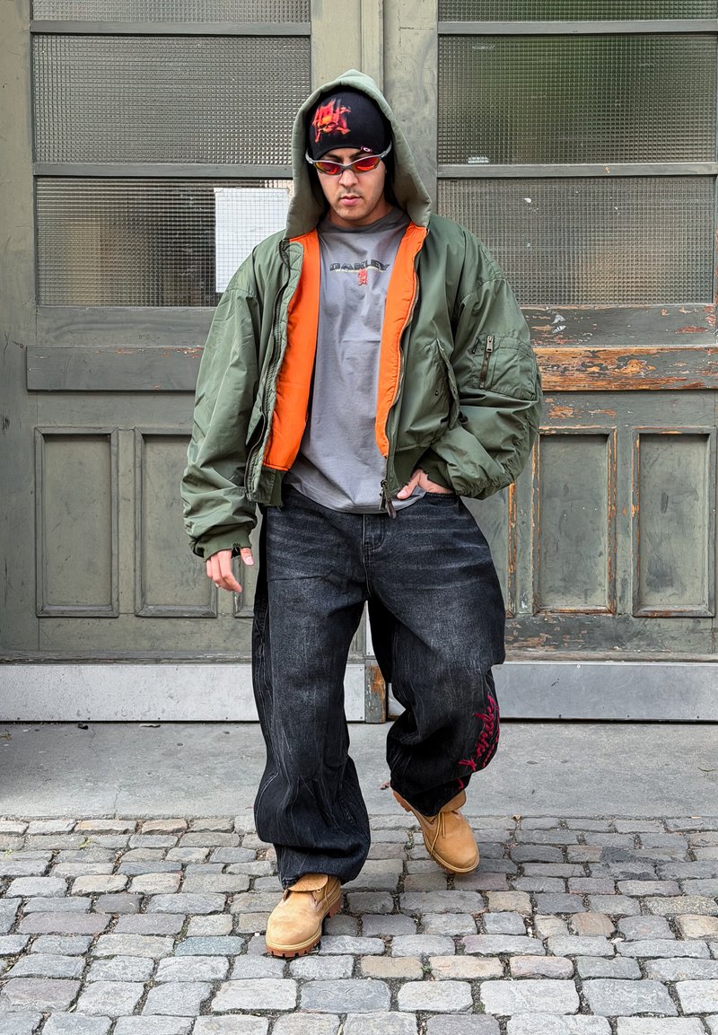 Ung mand i en stor grøn jakke med orange foer, sorte løstsiddende jeans, tanfarvede støvler, røde solbriller og en sort hue går på brosten.