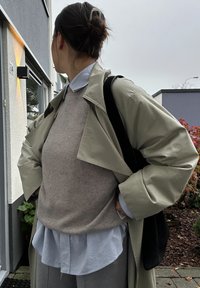 Καμτσένο κιτρινοπράσινο παλτό που είναι layered πάνω από ένα μπεζ πουλόβερ και ένα μπλε πουκάμισο, συνδυασμένο με γκρι παντελόνι. Μαύρη τσάντα κρεμασμένη στον ώμο, με τα μαλλιά σε κοτσίδα.