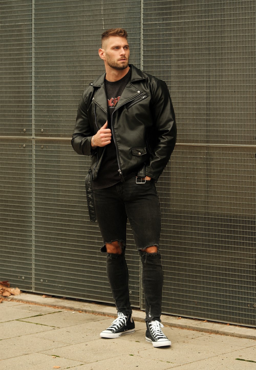 Casaco de couro negro de motociclista com detalhes em prata, t-shirt preta, jeans rasgados pretos e ténis pretos com solas brancas, apoiado contra um fundo de metal.