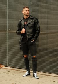 Casaco de couro negro de motociclista com detalhes em prata, t-shirt preta, jeans rasgados pretos e ténis pretos com solas brancas, apoiado contra um fundo de metal.