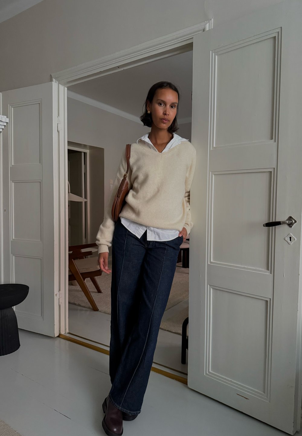 Maglione a maglia crema sopra una camicia bianca con colletto, abbinato a pantaloni in denim scuro. Borsa marrone, in piedi in una porta con un pavimento chiaro.