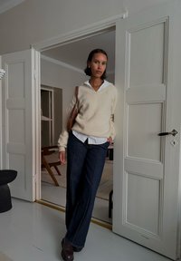 Maglione a maglia crema sopra una camicia bianca con colletto, abbinato a pantaloni in denim scuro. Borsa marrone, in piedi in una porta con un pavimento chiaro.