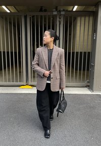 Brūns pleda žakete pār pelēku džemperi, melnas bikses un potīšu zābaki. Tur rokā nelielu melnu somiņu. Fona attēlā redzama aplenkta teritorija.