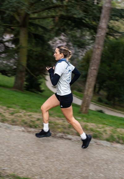 Corredor con una camiseta gris con un diseño azul, pantalones cortos negros y zapatillas deportivas negras, moviéndose rápidamente por un sendero de grava con vegetación cercana.