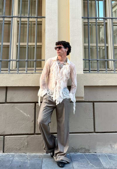 Blusa blanca fluida y transparente con detalles de volantes y un borde de encaje, combinada con pantalones de raya diplomática de pierna ancha en color marrón claro y zapatos negros.