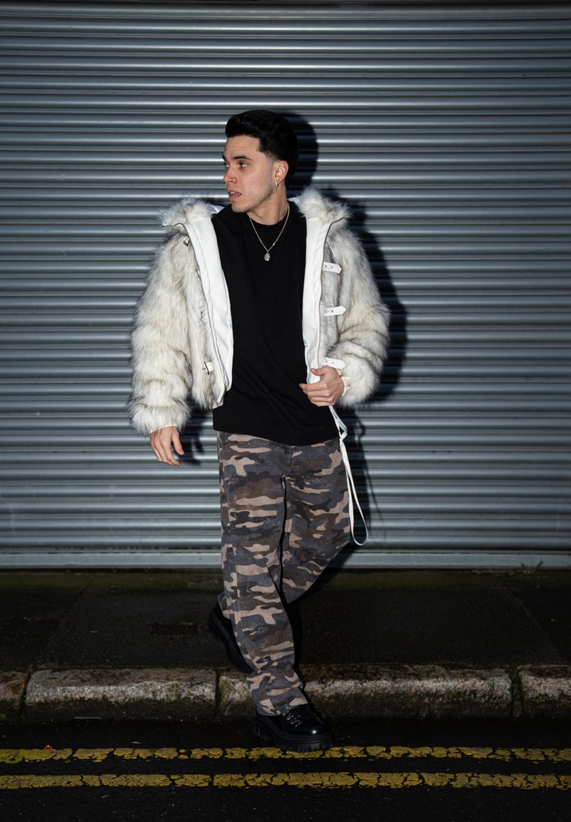 Jeune homme portant une veste en fourrure blanche, une chemise noire et un pantalon camouflage, debout dans une rue devant un rideau métallique.