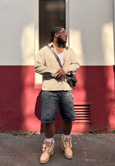 Camisa de cuadros beige y blanca de manga larga sobre una camiseta blanca sin mangas, pantalones cortos de mezclilla azules, botas beige y un bolso cruzado negro.