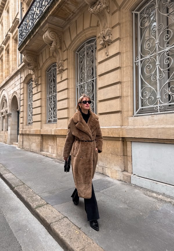 Brauner Kunstpelzmantel mit einem taillierten Knopfverschluss, doppelter Knopfleiste, schwarzem Rollkragenpullover und weit geschnittenen dunklen Jeans. Sonnenbrille und kleine Handtasche.