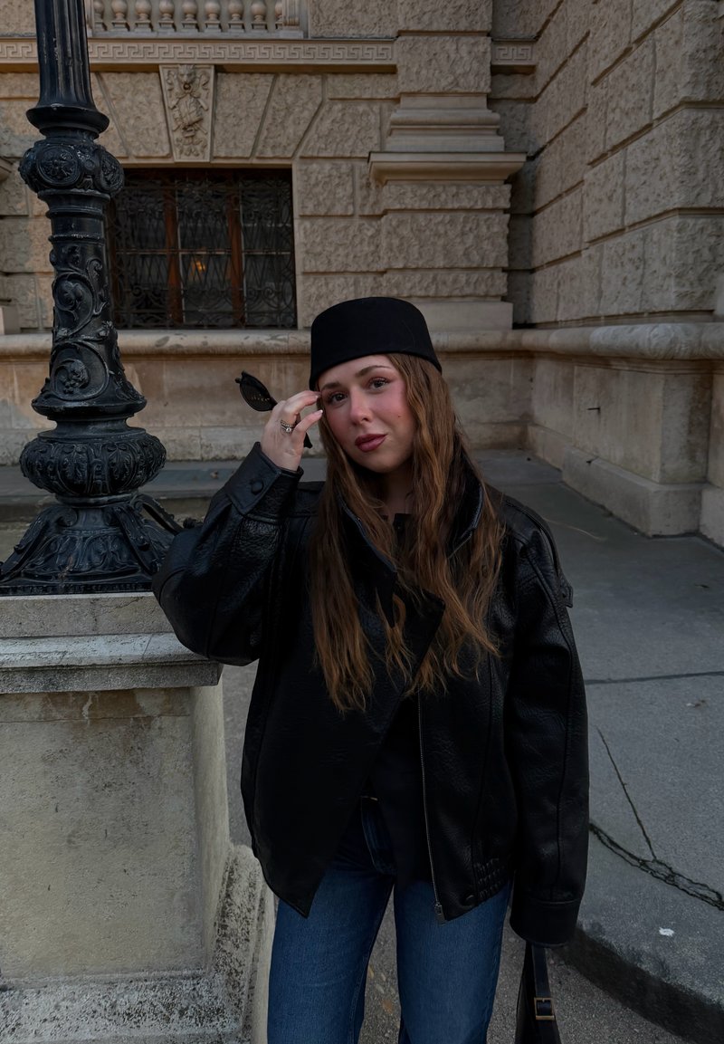 Jeune femme aux longs cheveux bruns porte une veste en cuir noire et un chapeau noir, tenant des lunettes de soleil près d'un lampadaire décoratif à l'extérieur d'un bâtiment en pierre.