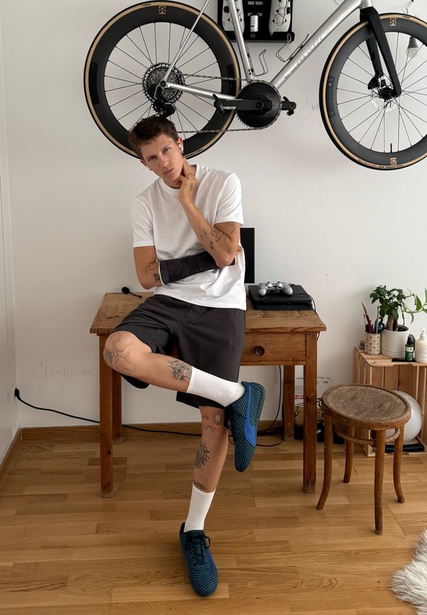 Mann mit Gipsarm und Tätowierungen sitzt an einem hölzernen Schreibtisch unter einem hängenden Fahrrad in einem Raum mit Holzboden und Pflanzen.