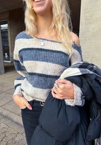 Pulover tricotat cu dungi orizontale gri, crem și bleumarin; design pe umeri descoperiți. Ține o geacă puff neagră cu model în carouri.