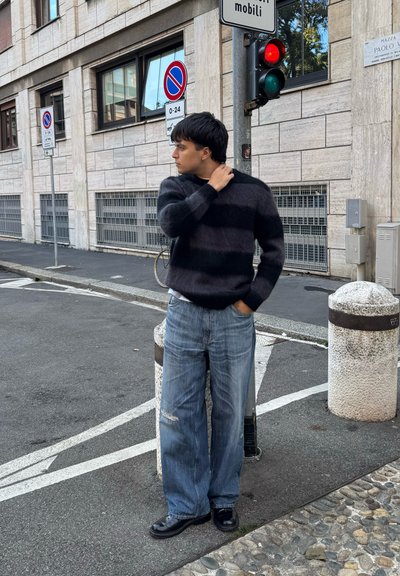 Suéter de punto a rayas negras y grises, jeans azules holgados con detalles desgastados y zapatos negros. De pie en una esquina de la calle cerca de señales de tráfico.