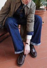 Καφέ δερμάτινα παπούτσια με ελαστικές σόλες, μπλε τζιν σαλοπέτες φαρδιές, πράσινη και καφέ ριγέ γραβάτα και ένα ανοιχτό μπουφάν. Φθαρμένες άκρες παντελονιού.