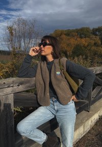 Colete de fleece castanho por cima de um suéter escuro, combinado com jeans azul claro. Os acessórios incluem óculos de sol e uma bolsa verde. Fundo natural.