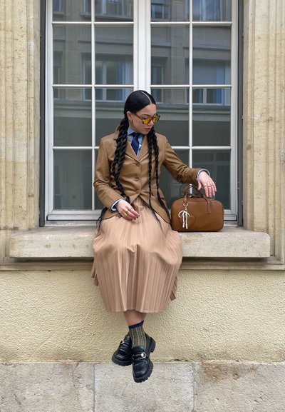 Blazer de cuero marrón sobre una camisa azul, combinado con una falda beige con pliegues y mocasines negros. Sostiene un bolso marrón con un charm.