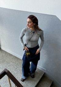 Chemise grise à manches longues, jupe en denim foncé superposée sur un jean large, béret marron, collier voyant, accessoire floral sur la poche de la jupe.