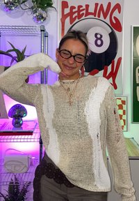 Ανοιχτό μπεζ πλεκτό πουλόβερ με υφή και φθαρμένες λεπτομέρειες, συνδυασμένο με στρωματωμένες αλυσίδες. Το φόντο περιλαμβάνει ζωντανή διακόσμηση και φυτά.