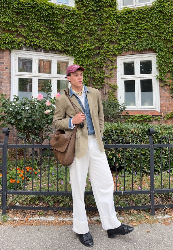 Jeune homme portant un blazer beige, un pantalon blanc et une casquette bordeaux, se tient près d'une clôture noire devant un bâtiment en briques couvert de lierre et de fleurs.