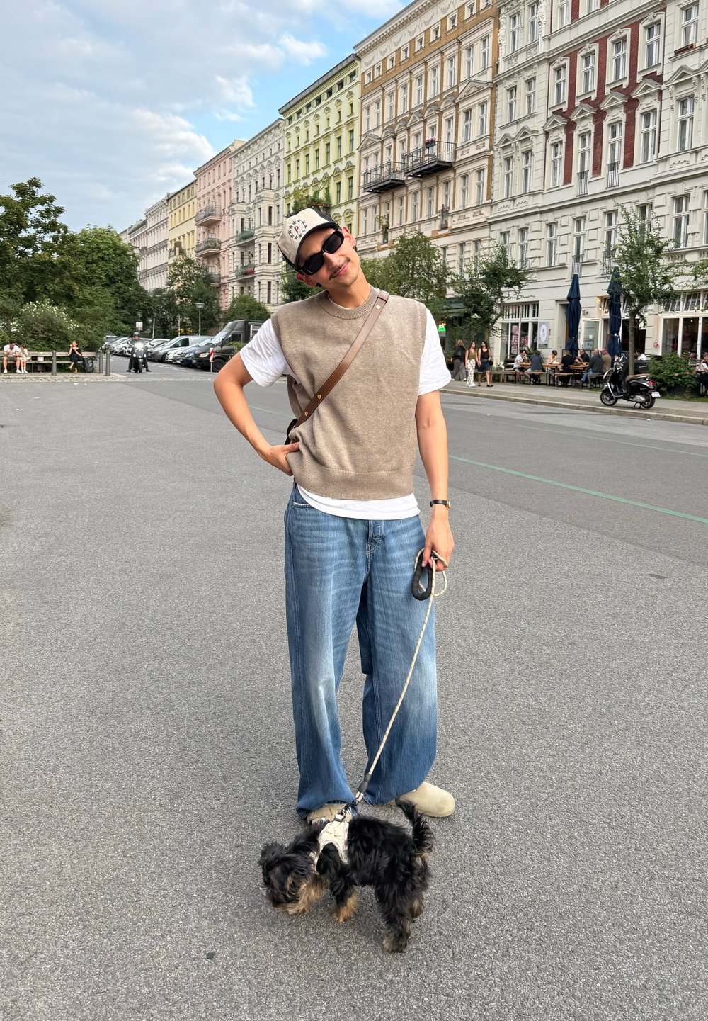 Henkilö, joka käyttää aurinkolaseja, lippistä, vaaleanbeigeä liiviä ja farkkuja, seisoo pienen mustan koiran kanssa hihnassa urbaanilla kadulla, jossa on korkeita rakennuksia ja ulkoistumista.