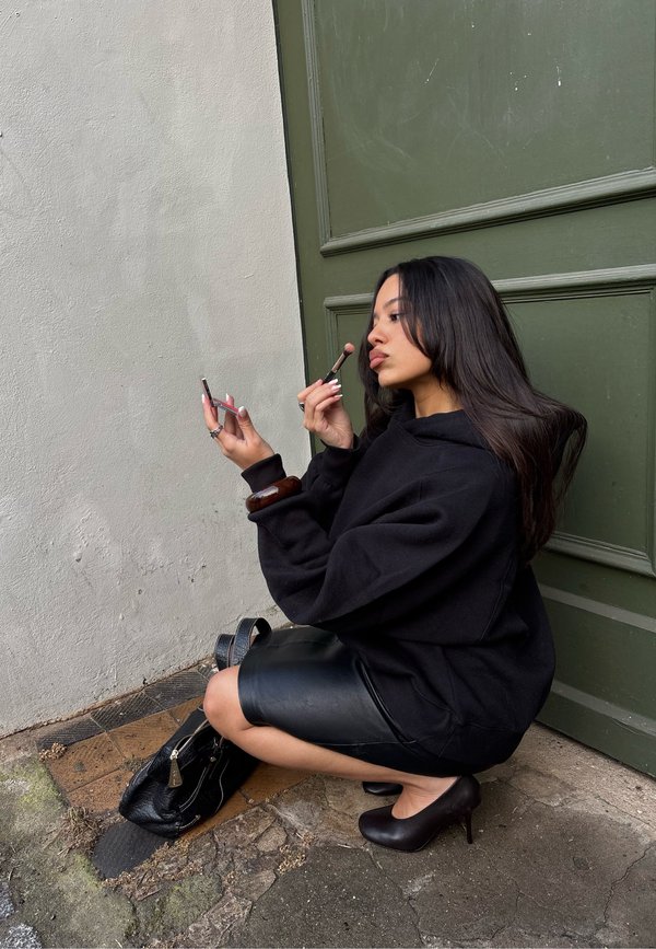 Vrouw in een zwarte hoodie en leren rok hurkt bij een groene deur, terwijl ze make-up aanbrengt met een compact spiegel en een kwast.