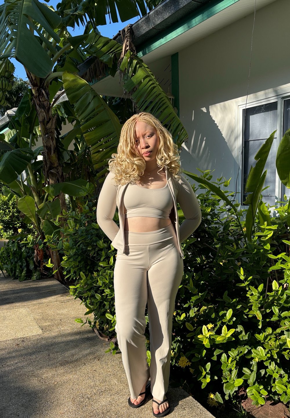 Beige two-piece outfit made of smooth fabric, featuring a fitted crop top and high-waisted trousers. Natural surroundings include green foliage.