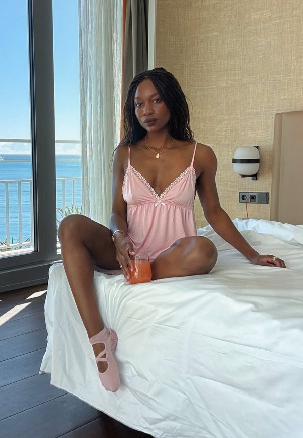 Femme en pyjama rose et chaussettes assise en tailleur sur un lit, tenant un verre de jus d'orange, avec vue sur la mer à travers une grande fenêtre.
