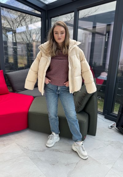 Chaqueta de plumas beige con cuello alto, suéter marrón, vaqueros de colour azul claro y zapatillas blancas gruesas, de pie contra un fondo de cristal.