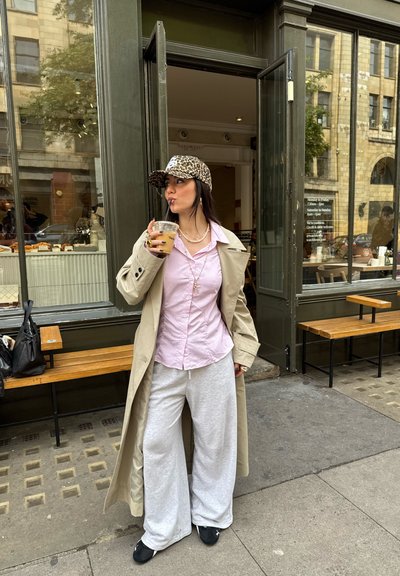 Trench coat beige, camisa de botón rosa claro, pantalones de chándal grises de pierna ancha, gorra de estampado de leopardo, sosteniendo una bebida, de pie fuera de un café.