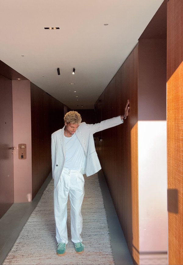 Persona con capelli ricci che indossa un blazer bianco a righe, pantaloni bianchi e scarpe verdi appoggiata contro una parete di legno nel corridoio.