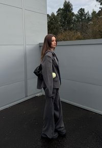 Jeune femme en costume gris foncé surdimensionné et chaussures à plateforme, debout en extérieur contre un mur gris, avec des arbres en arrière-plan.