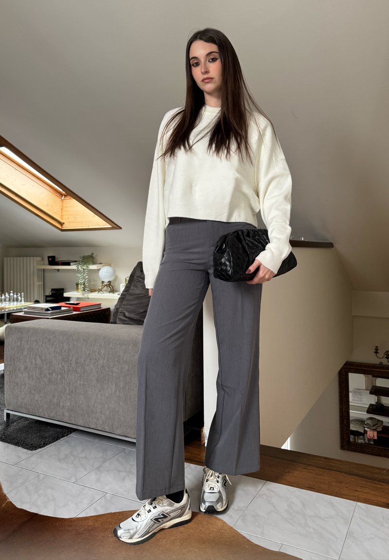 Jeune femme debout à l'intérieur portant un pull blanc, un pantalon gris à jambes larges, des baskets argentées et tenant une pochette noire.