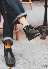 Black leather shoes with laces, a round toe, and a textured sole. Yellow ribbed socks featuring logo detailing. Rolled denim trousers with contrast stitching.