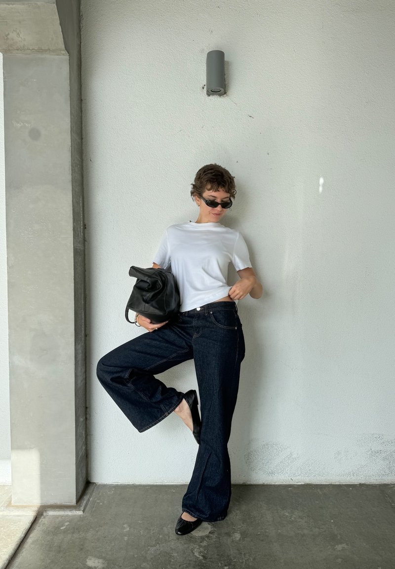 Camisa branca de manga curta, jeans de perna larga azul escuro, sabrinas de couro pretas e uma bolsa preta, posando contra uma parede cinza clara.
