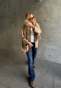 Rjava suedna jakna s frinči ob bele srajce, modri flare džinsih in rjavih gležnjarjih. Rjava torba čez ramo in temna sončna očala zaokrožita videz.