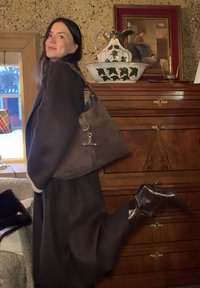 Χαμογελαστή γυναίκα με σκούρο παλτό και μπότες σηκώνει το ένα πόδι, κρατώντας μια μεγάλη καφέ σουέντ τσάντα, όρθια δίπλα σε ξύλινο κομοδίνο με διακοσμητικό κανάτι.