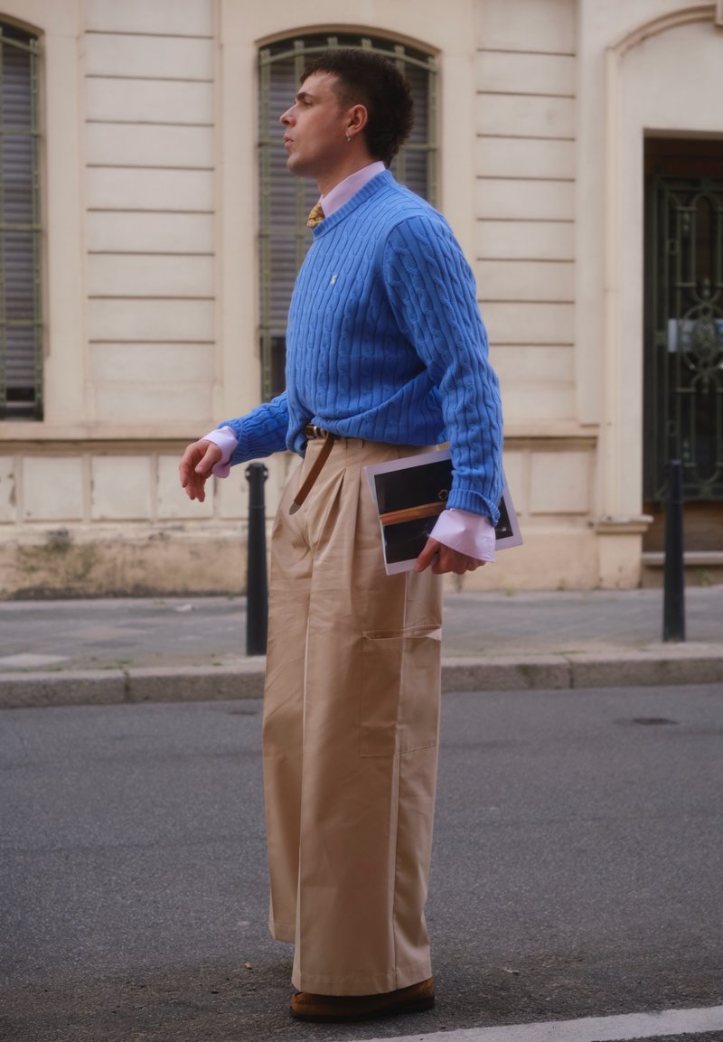 Man walking on street wearing a blue sweater, beige wide pants, holding a tablet, with historic building and windows in the background.
