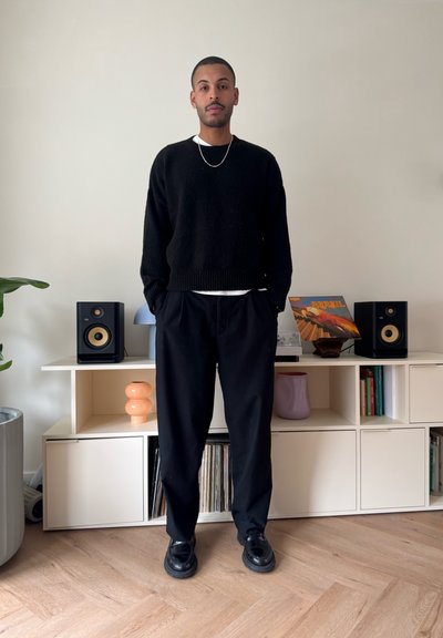 Hombre con suéter negro y pantalones, de pie con las manos en los bolsillos, en una habitación moderna con altavoces, discos de vinilo y jarrones decorativos en estantes.