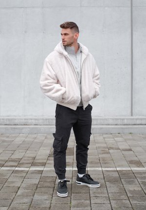 Hombre de pie al aire libre con una sudadera con capucha gris claro, chaqueta blanca y esponjosa, pantalones cargo negros y zapatillas negras sobre una superficie de baldosas.