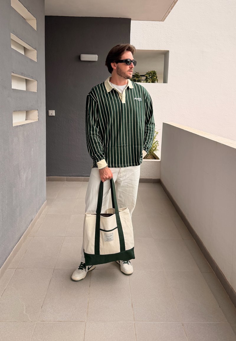 Homme portant une chemise à manches longues rayée, pantalon blanc, lunettes de soleil, tenant un grand sac fourre-tout blanc et vert, debout sur un balcon carrelé.
