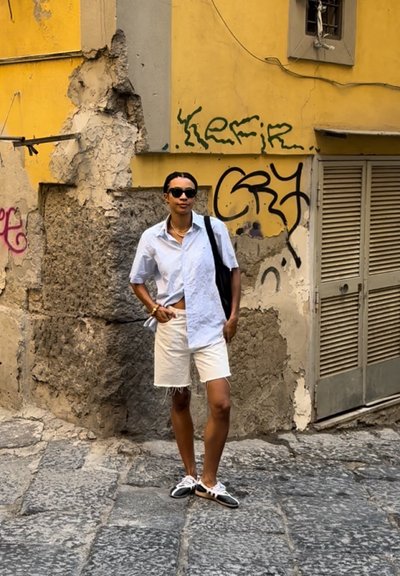 Camisa blanca de manga corta, pantalones cortos de color claro y zapatos negros con cordones blancos. De pie sobre adoquines, fondo de graffiti en una pared amarilla.