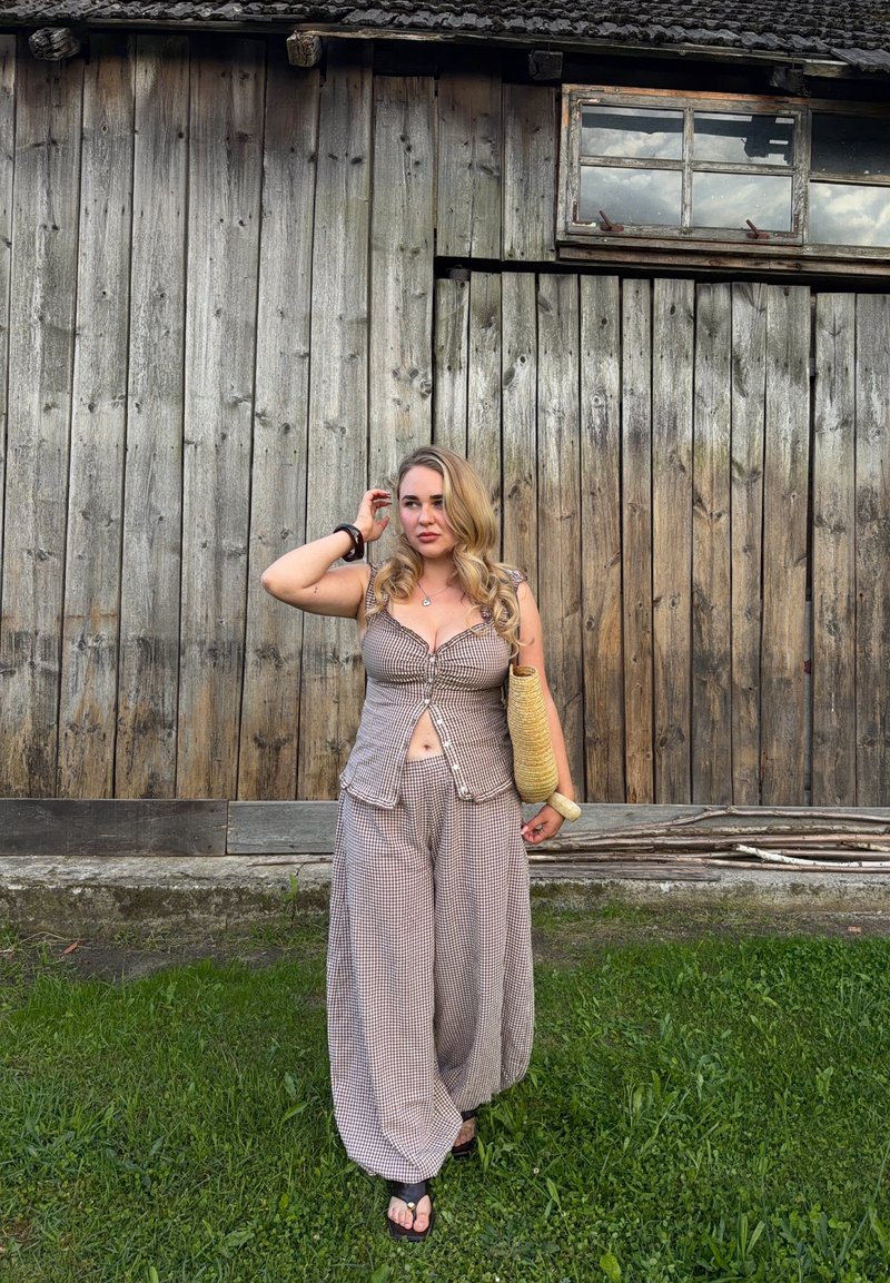 Žena s dlouhými blond vlasy stojí na trávě před povětrnostmi oprýskanou dřevěnou stodolu, má na sobě kostkovaný top a široké kalhoty a drží pletenou tašku.