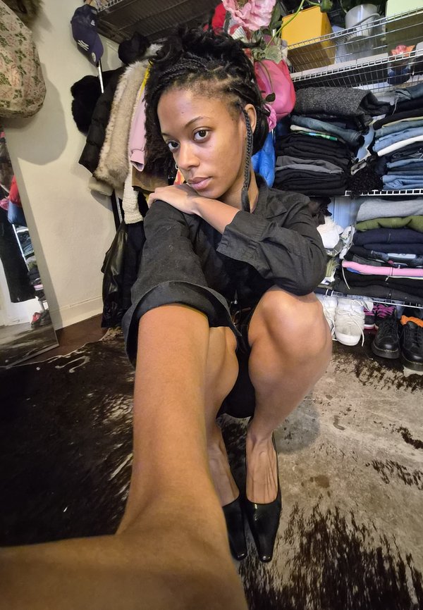 Melns pogu krekls, ceļos lūgšanas poza, melni augstpapēžu apavi, fakturēts fons ar drēbēm uz plauktiem, silta apgaismojuma, minimāla nekārtība.