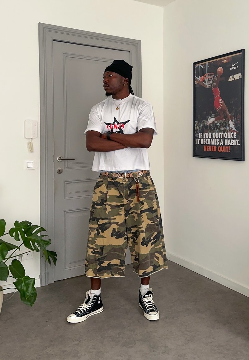 Mann trägt weißes T-Shirt, Camouflage-Shorts, schwarze Mütze und Turnschuhe und steht mit verschränkten Armen in einem Raum mit einem Basketball-Motivationsposter.
